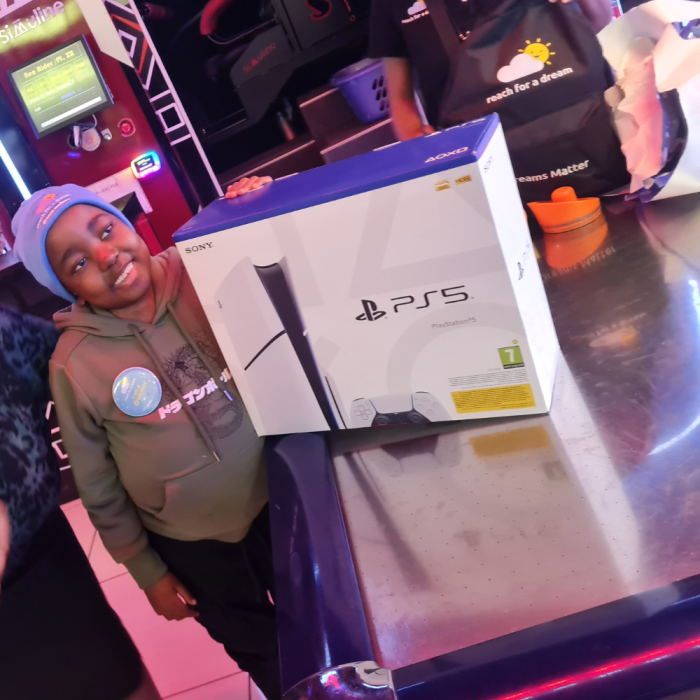 Image of a smiling young child at a games arcade with an unopened PS5 Box with a Reach for a Dream representative in the background