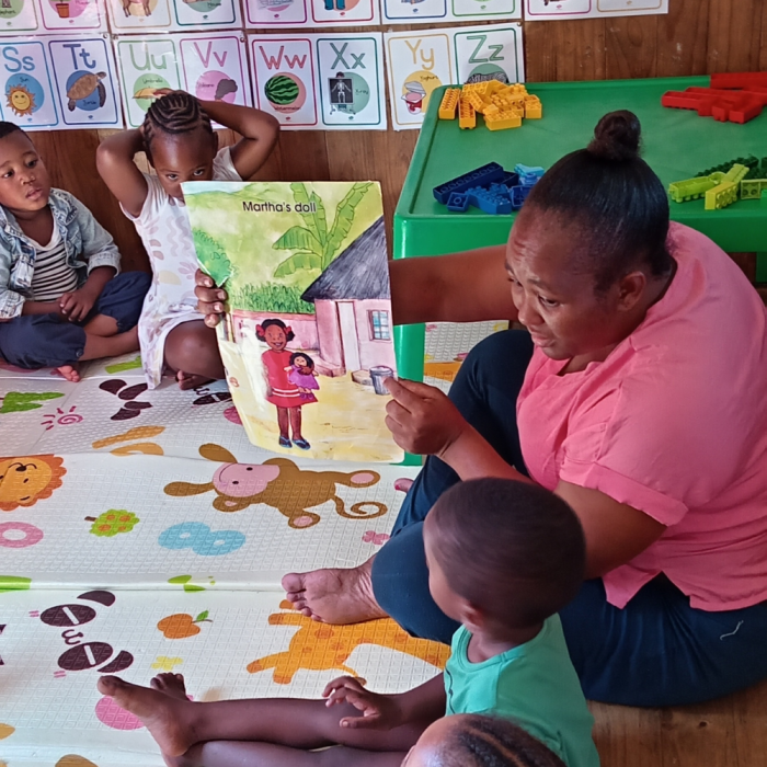 Image of an ECD teacher showing a story book to 4 children on the mat in class
