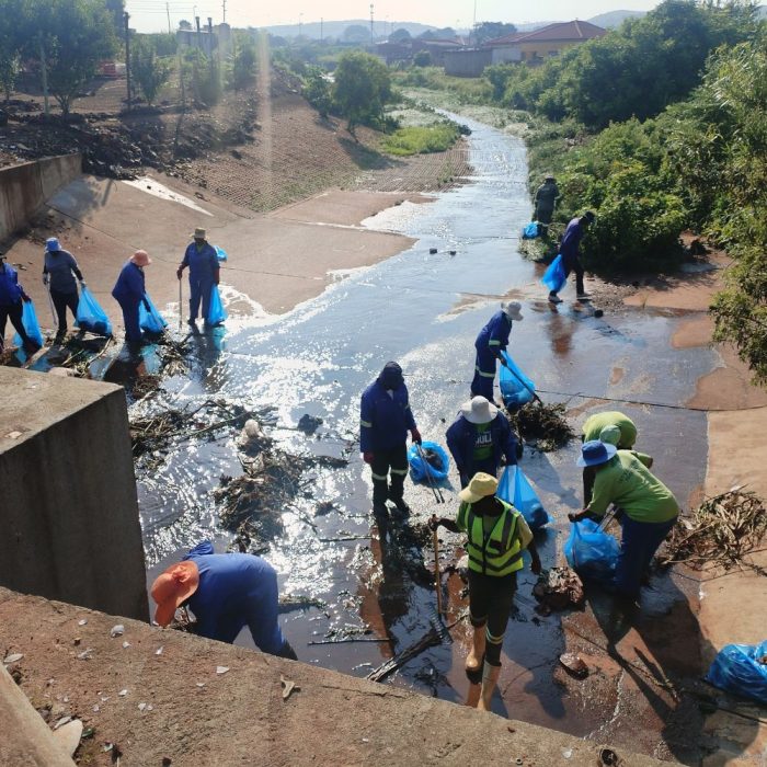 Image of 13 people wearing hats, overalls and gloves cleaning up the environment at a big drain under a bridge with brooms and plastic bags