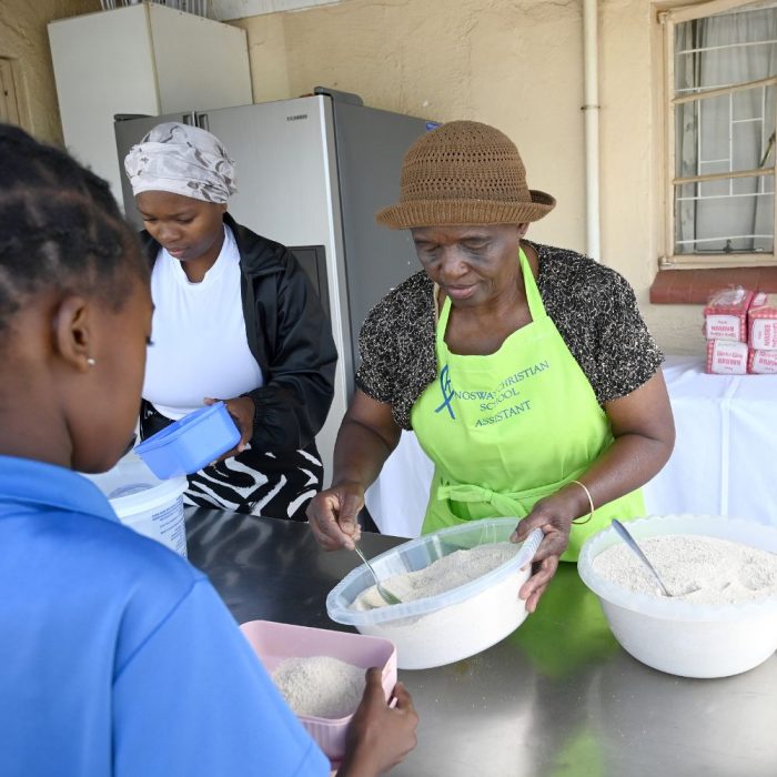 Image of two ladies scooping dry maize meal into lunchbox containers held by children in a line