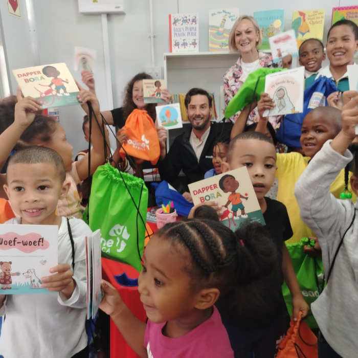 Image of 11 young children holding up children's books and smiling in a library with their 3 teachers