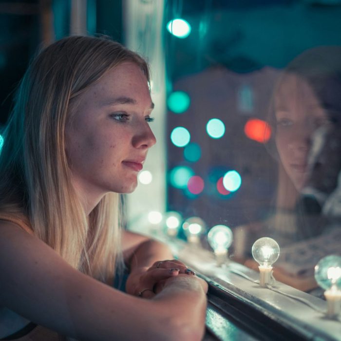 Image of a young, blonde lady looking out a window framed by lights at night